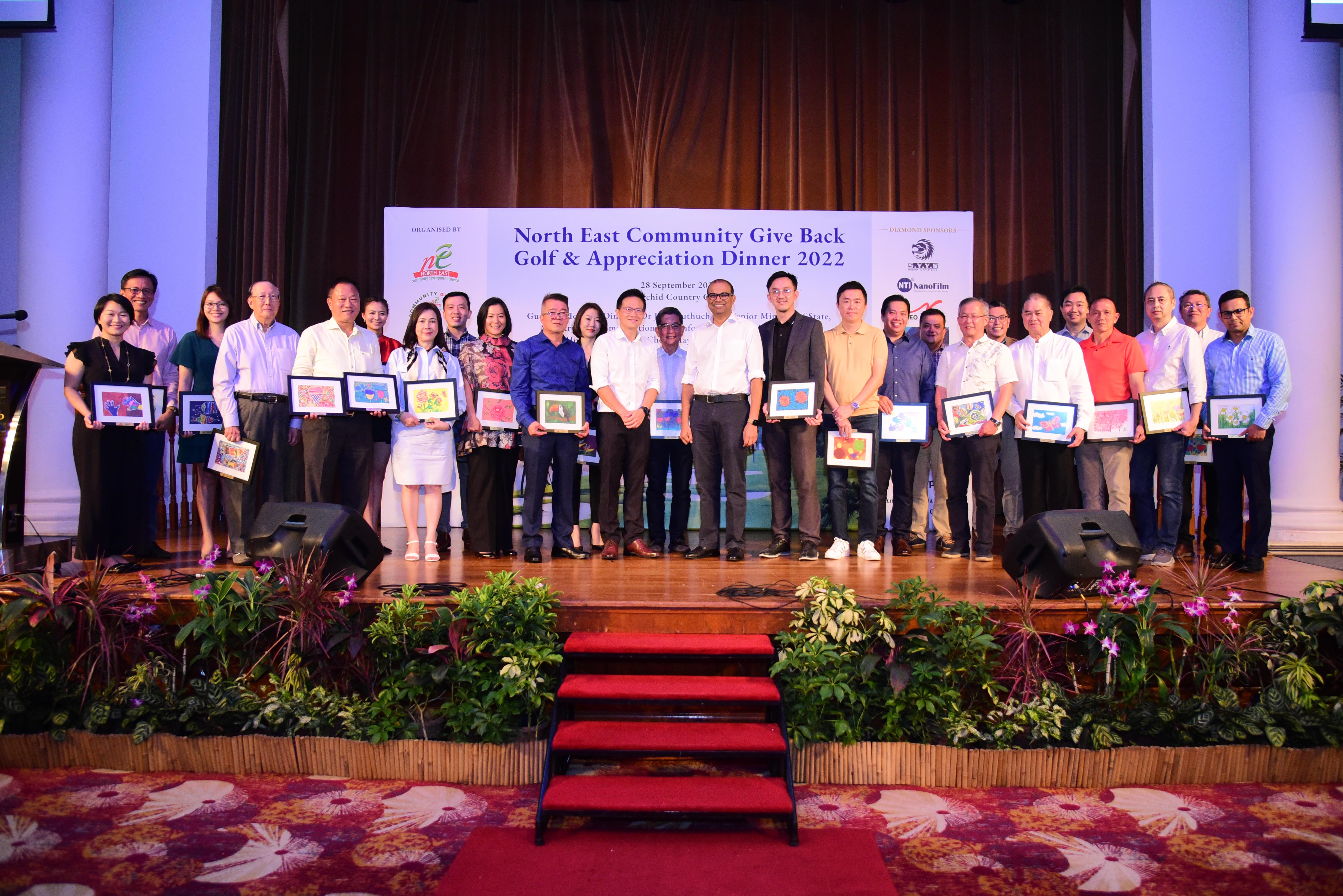 Group photo on stage at a community appreciation dinner, with guests and organisers holding framed artworks during the North East Community Give Back Golf & Appreciation Dinner 2022.